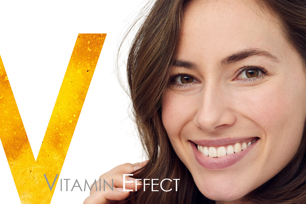Porträt einer lächelnden Frau mit natürlicher, strahlender Haut vor einem weißen Hintergrund mit  orange-gelben „V“. Symbolbild für den „Vitamin Effect“ – Energie, Vitalität und Leuchtkraft der Haut durch vitaminreiche Pflege.